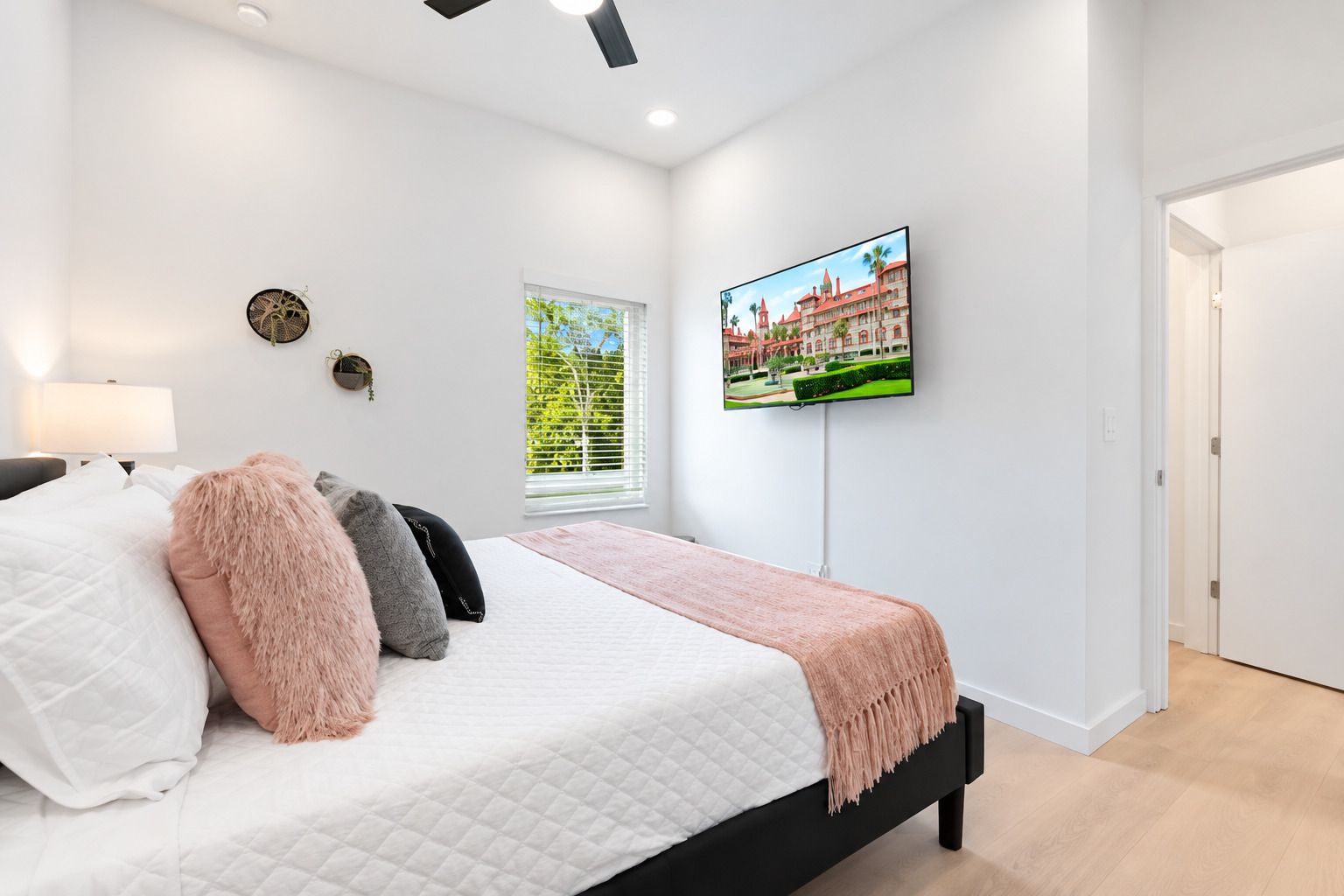 A bedroom with a bed, a TV mounted on the wall, a window with blinds, and a door.