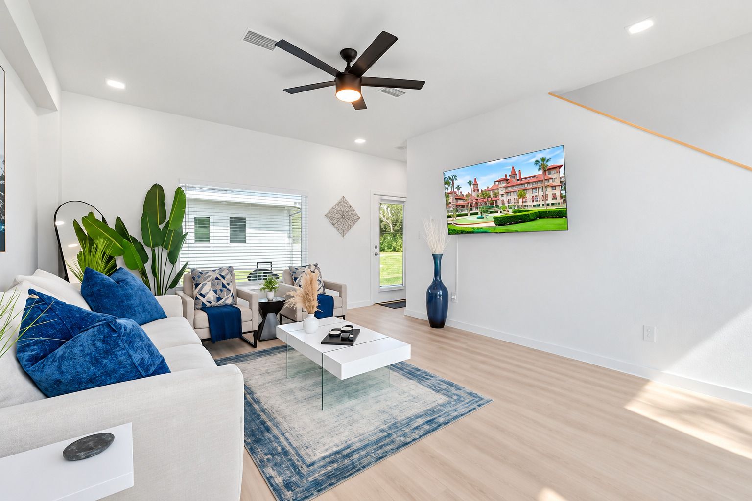 A modern living room with a white sofa, blue cushions, a coffee table, a TV on the wall, and a ceiling fan.