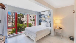 A modern hotel room with a large bed, a nightstand, a lamp, and a view of the city through large windows. - Red Door Inn (San Juan)