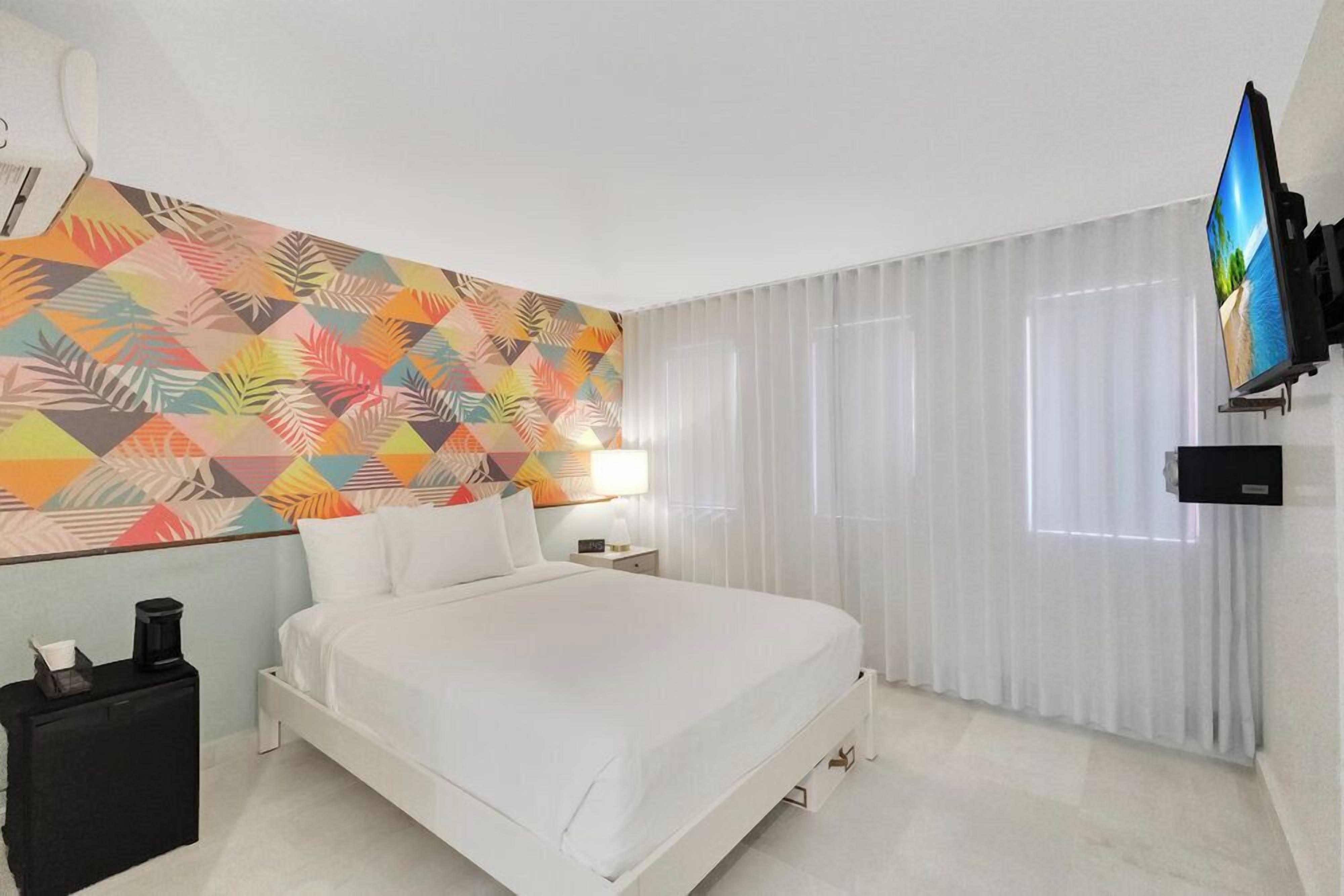 A hotel room with a bed, a TV mounted on the wall, a nightstand, and a colorful geometric-patterned wall.