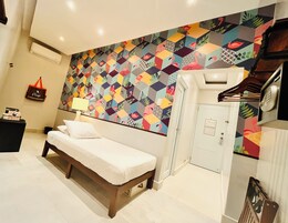 Habitación de hotel con cama, aire acondicionado de pared, mesita de noche con lámpara, armario y pared con un llamativo diseño geométrico.