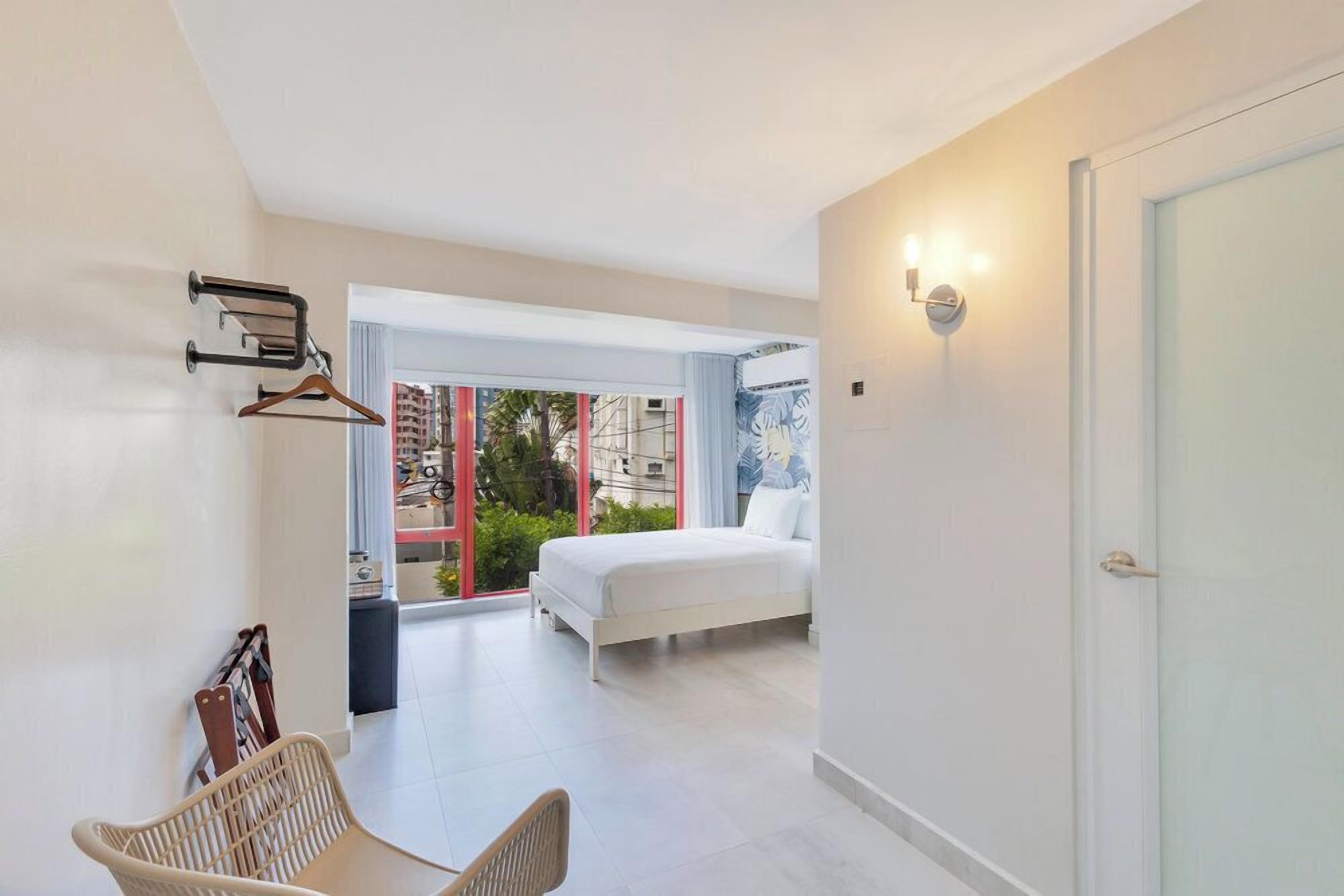 A modern hotel room with a bed, a chair, a hanging coat rack, and a view of the city through large windows.