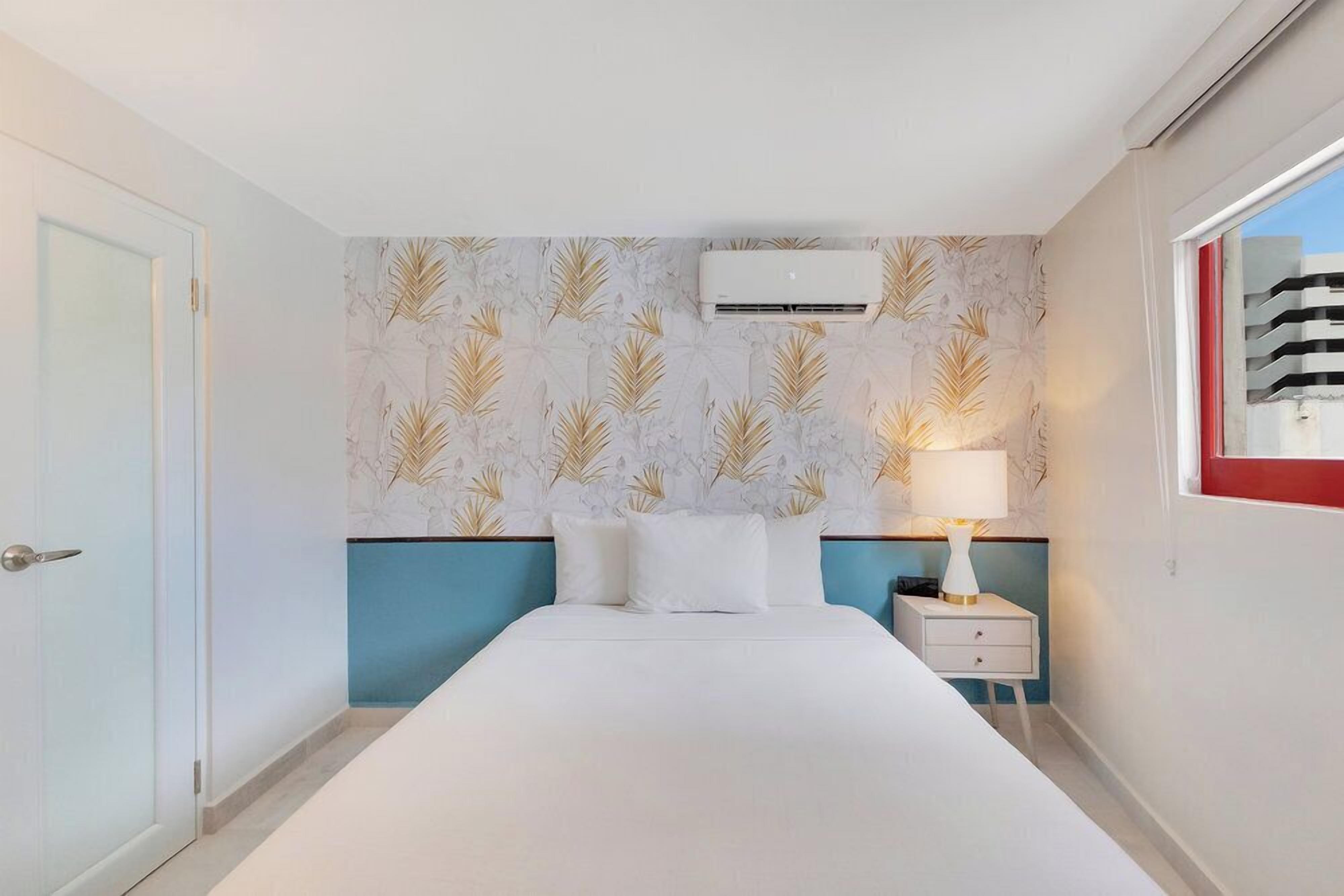 A bedroom with a bed, white walls, a patterned wallpaper, an air conditioning unit, a bedside table with a lamp, and a window with a red frame.