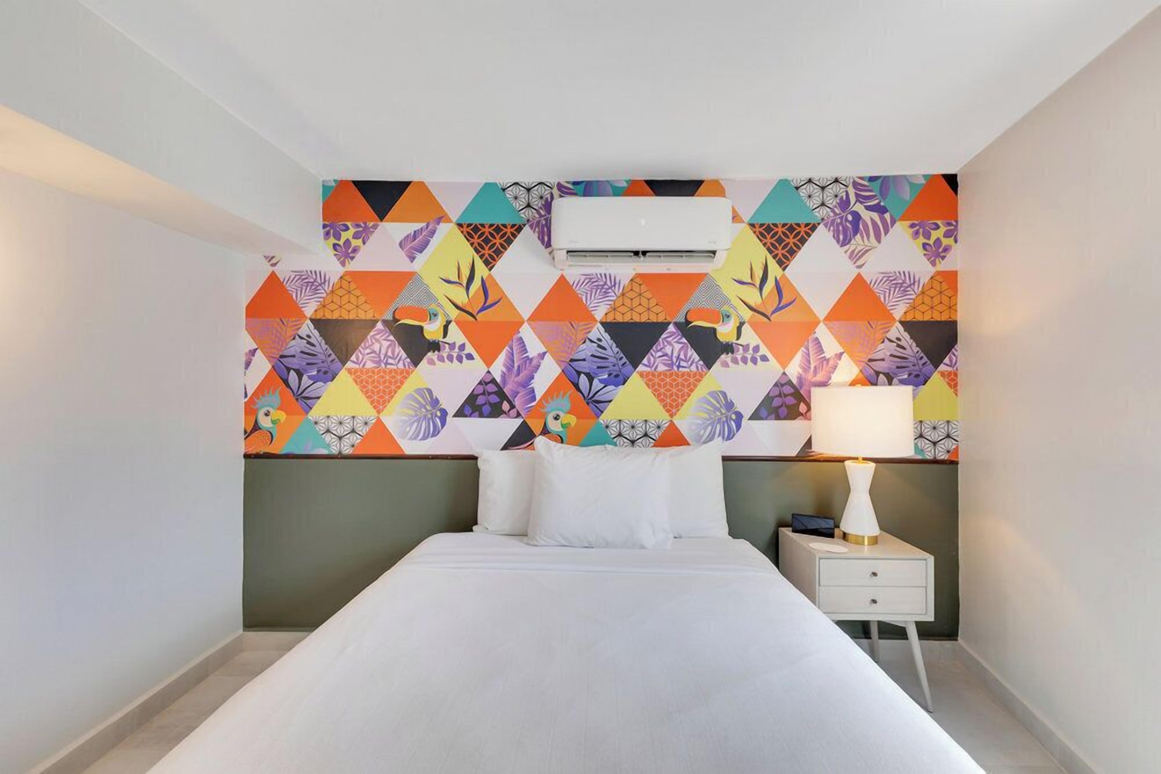 Een hotelkamer met een bed, een nachtkastje met een lamp, en een wand met een kleurrijk geometrisch patroon.