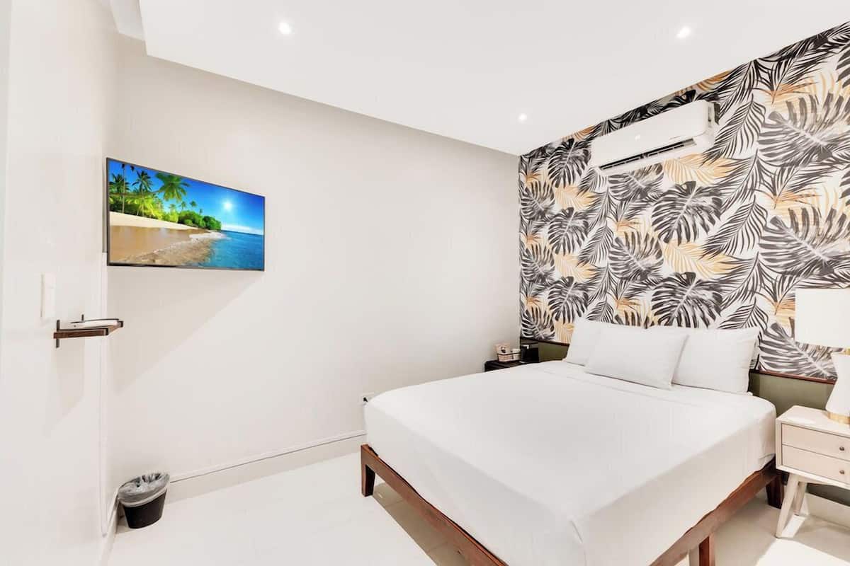 A bedroom with a bed, a nightstand, a wall-mounted air conditioner, a trash bin, and a picture of a beach scene.