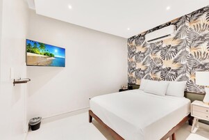 A bedroom with a bed, a nightstand, a wall-mounted air conditioner, a trash bin, and a picture of a beach scene.