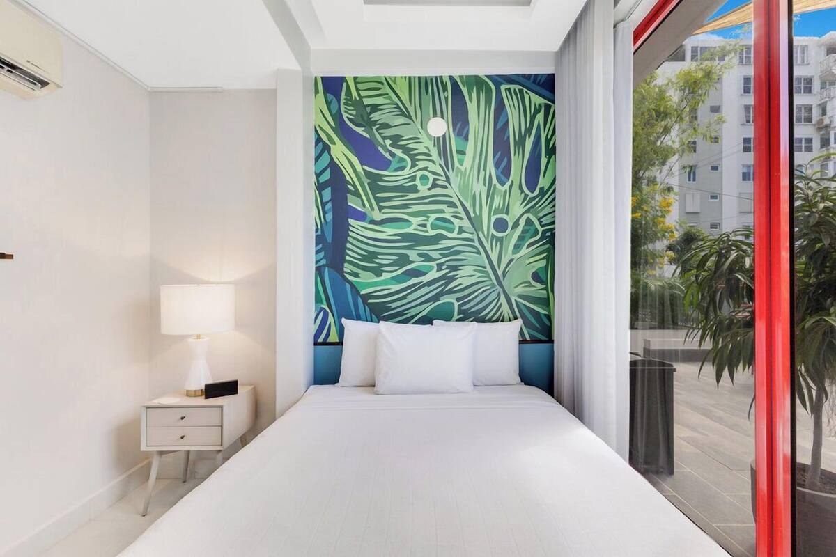 A hotel room with a large bed, a nightstand with a lamp, a colorful wall mural, and a view of the city through a large window.
