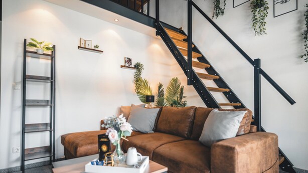 A modern living room with a brown leather sofa, a coffee table with remote controls, a ladder, and a staircase with wooden steps. - Vacation apartment in the countryside - with lake view, terrace & barbecue fun (Lohmen)