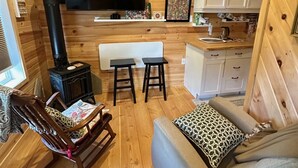 A cozy cabin interior with a wooden interior, a TV mounted on the wall, a fireplace, a sofa, a rocking chair, and a kitchen area with cabinets and a countertop.
