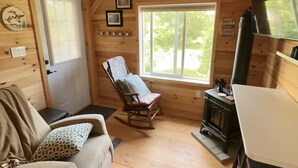 A cozy cabin interior with a wooden floor, a rocking chair, a sofa, a fireplace, and a large window providing natural light.