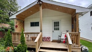 A wooden porch with steps leading to a door, surrounded by a garden with various plants and a small flag.
