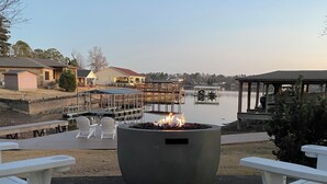 A patio with a fire pit, white Adirondack chairs, and a view of a lake with buildings in the background.