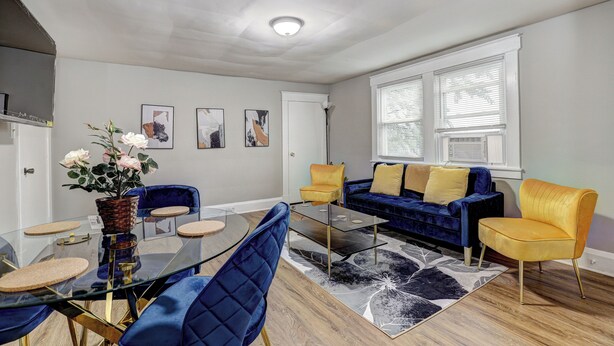 Una sala moderna con una mesa de centro de vidrio, sillas azules y amarillas, un sofá azul y un ventanal con persianas.