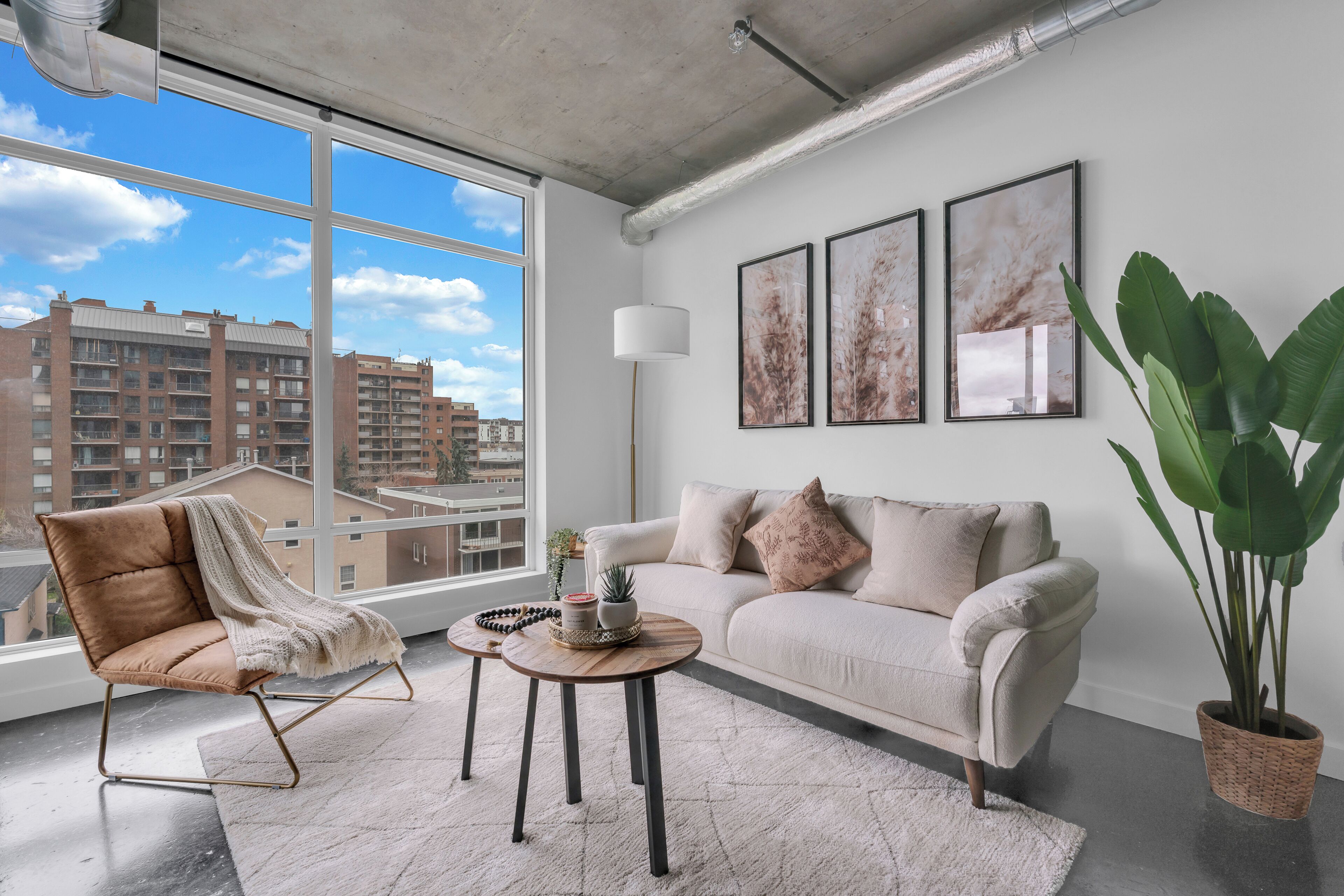 Een moderne woonkamer met een bank, fauteuil en salontafel, grote ramen met uitzicht op de stad, en drie ingelijste kunstwerken aan de muur.