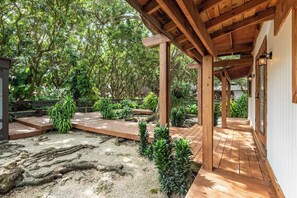 Eine Holzterrasse mit einer Pergola, umgeben von üppigem Grün und einer Sandfläche.