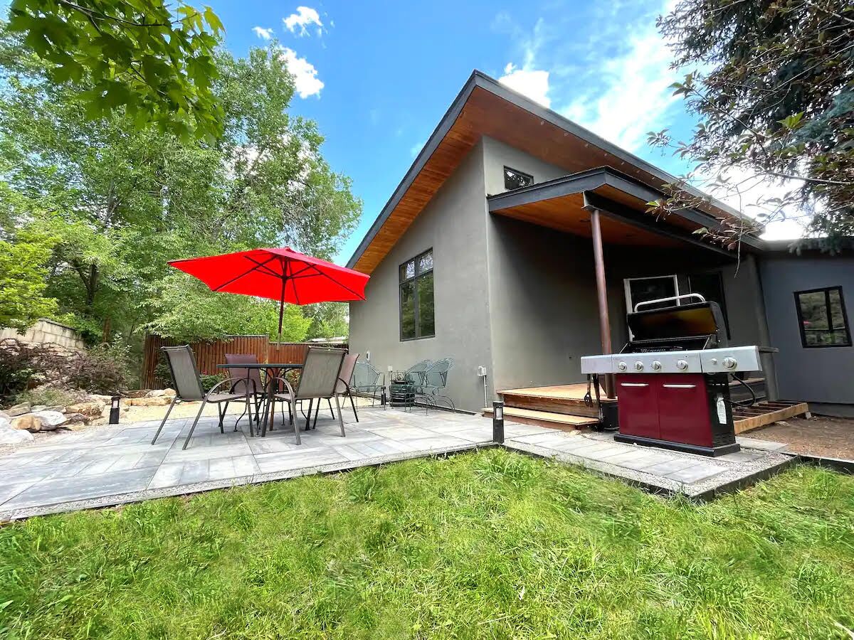 Une maison moderne dotée d’un patio comprenant une table et des chaises, un parasol rouge et un barbecue.