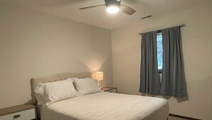 A bedroom with a bed, a nightstand, a lamp, a ceiling fan, and a window with curtains.