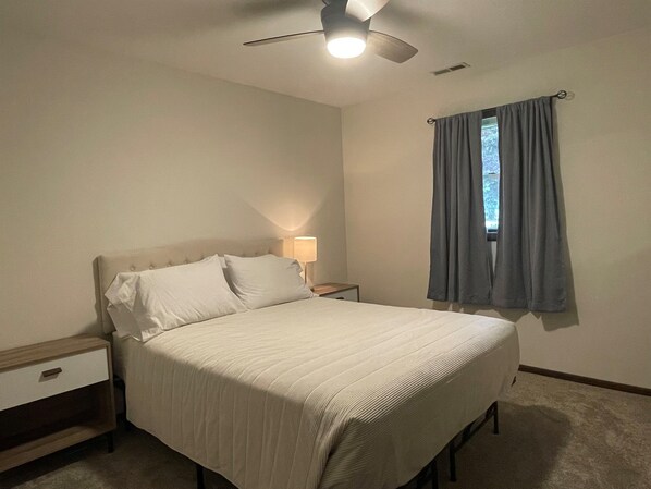 A bedroom with a bed, a nightstand, a lamp, a ceiling fan, and a window with curtains.