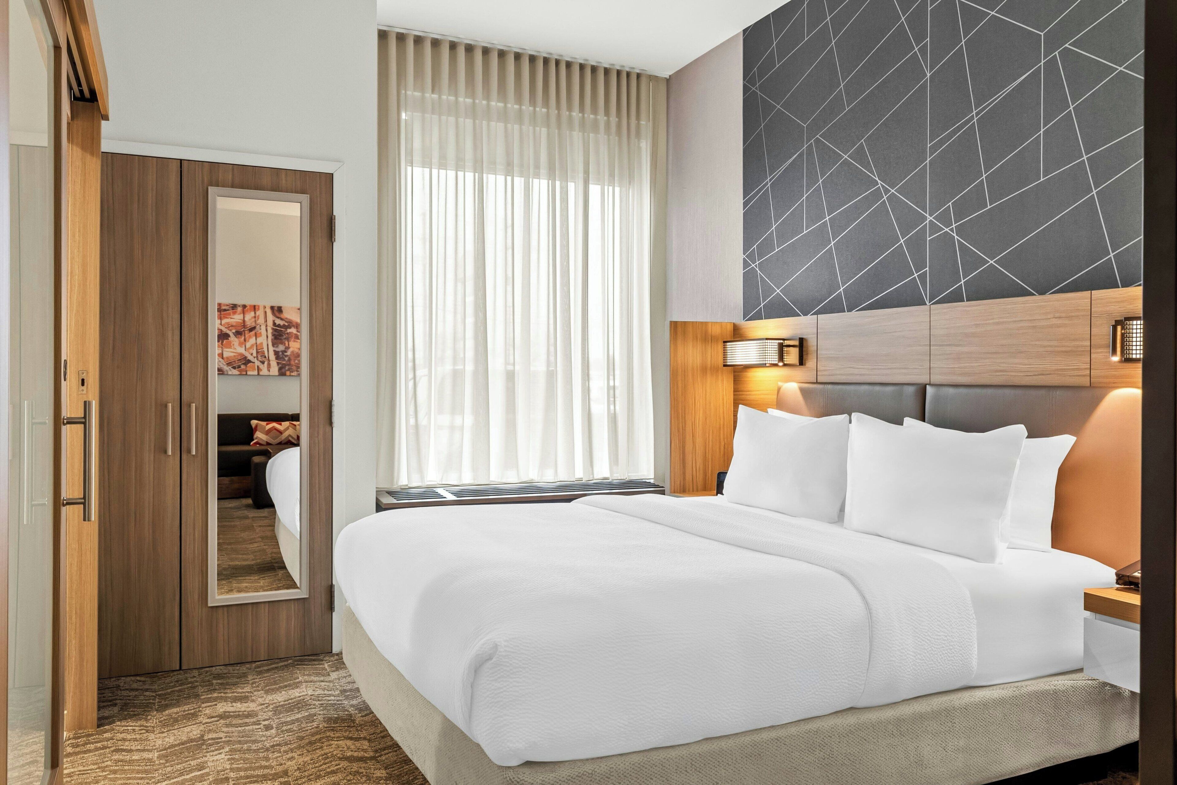 A hotel room with a large bed, a wooden wardrobe, a mirror, a window with sheer curtains, and a geometric-patterned wall.