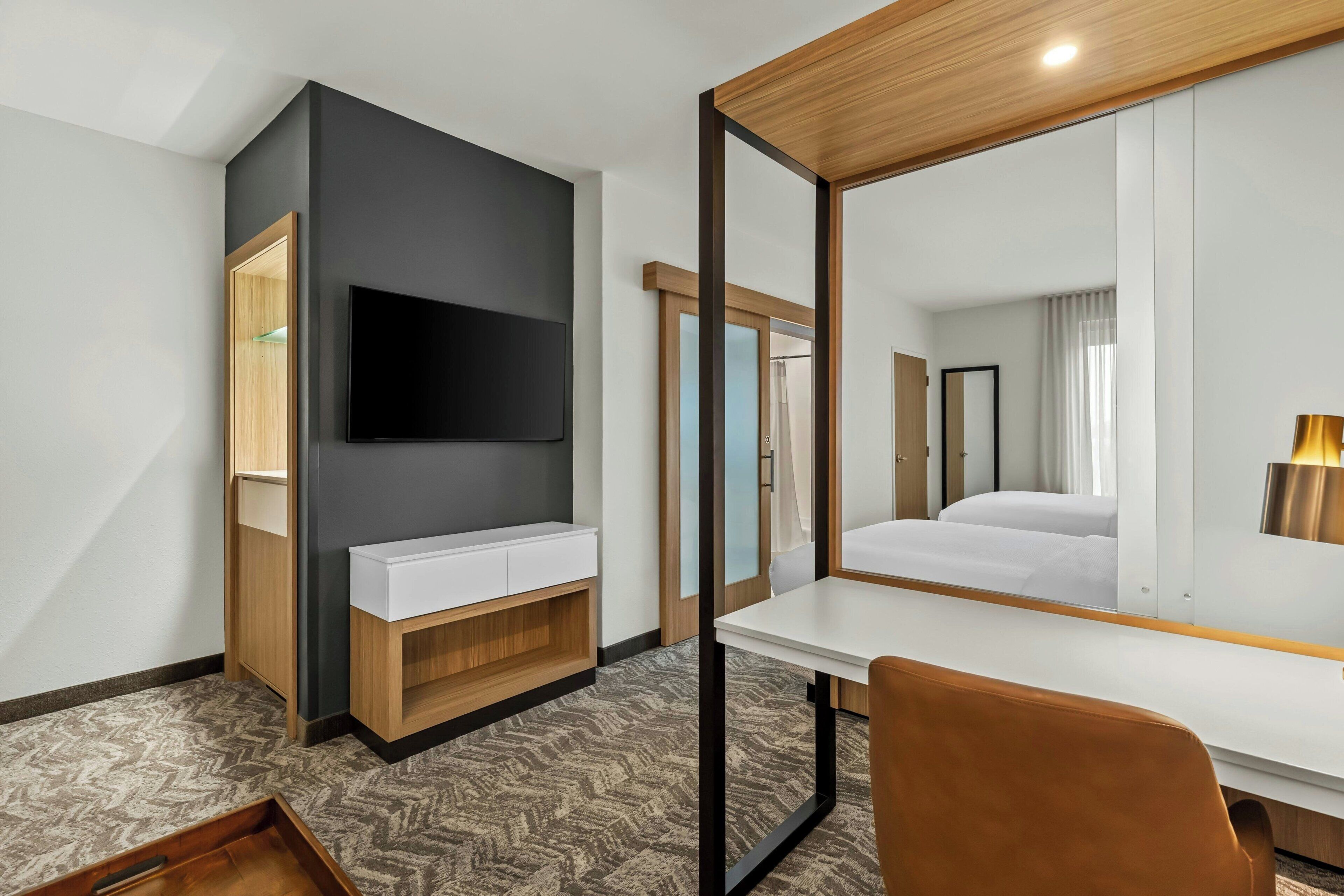Une chambre d’hôtel équipée d’un téléviseur à écran plat, d’un bureau blanc, d’une chaise marron, d’une armoire en bois et d’un grand miroir.