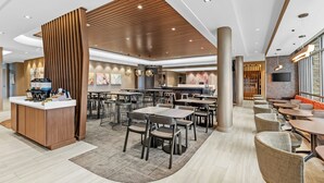 En modern hotellobby med en central receptionsdisk, flera sittgrupper och en stor TV-skärm.