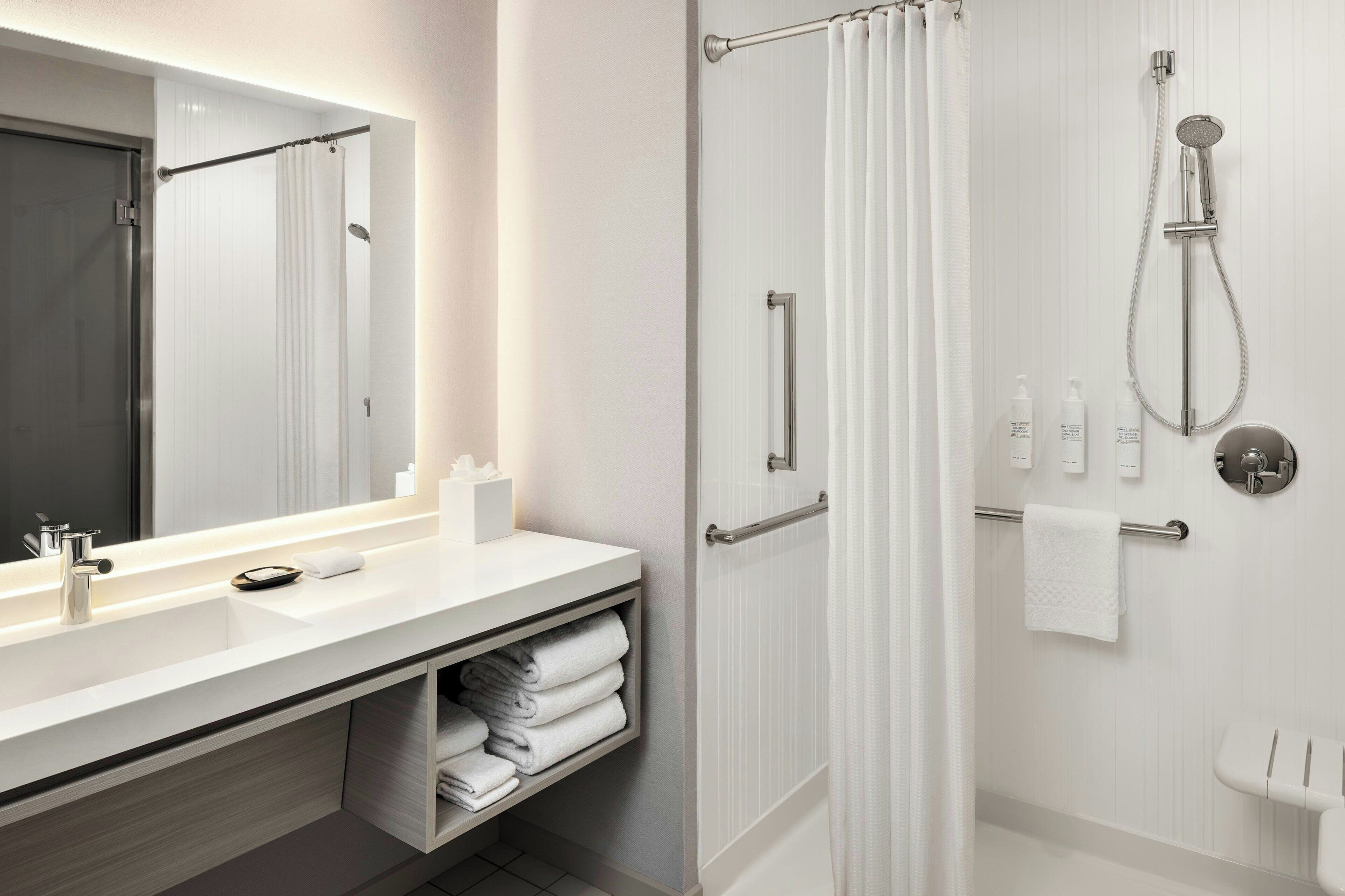 A modern bathroom with a large mirror, a shower with a handheld showerhead, a toilet, and a vanity with a sink and towel storage.