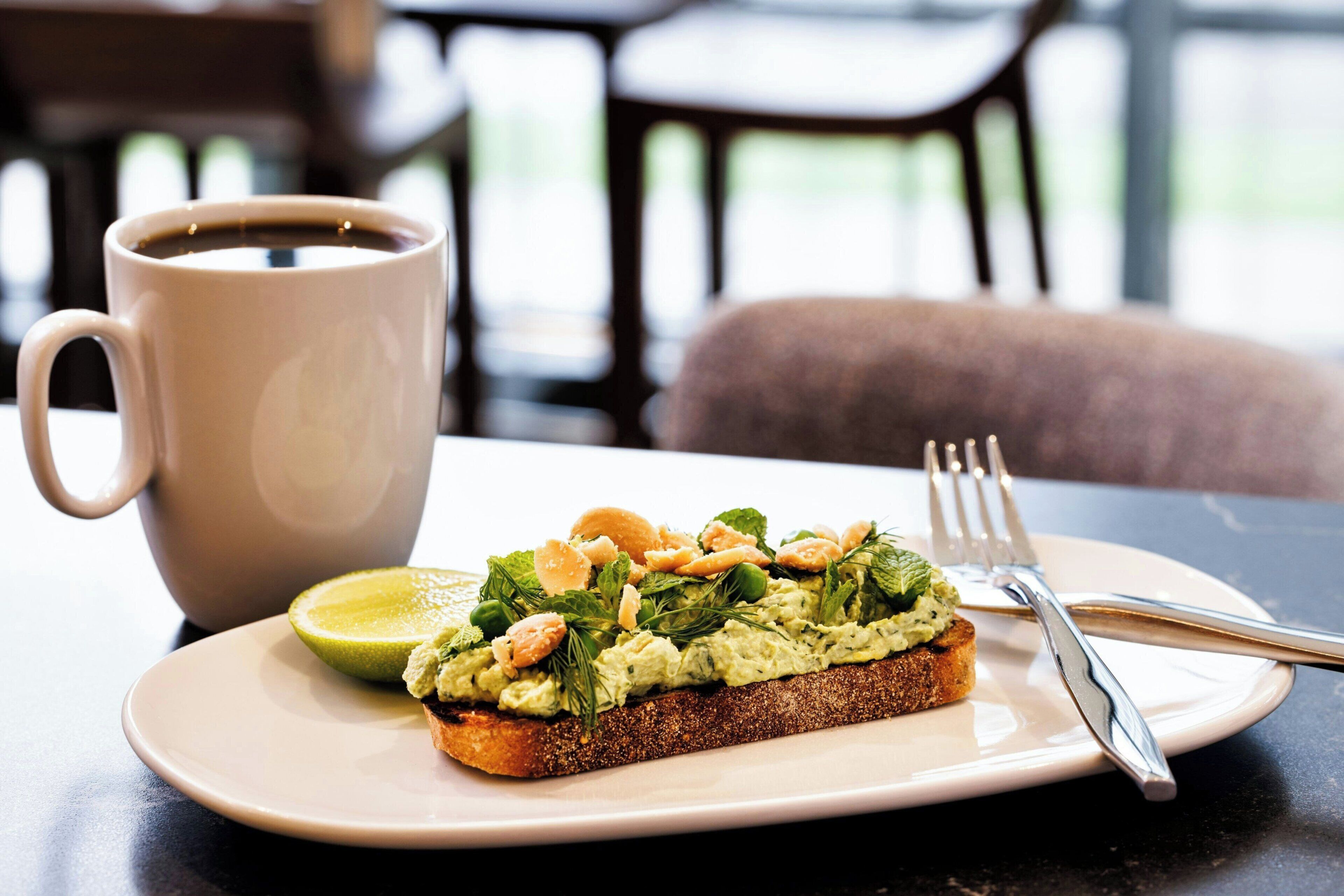 A slice of toast with avocado spread, a lime wedge, and nuts on a plate, accompanied by a coffee mug and cutlery on a table.