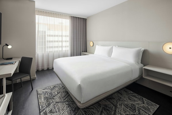 Habitación de hotel con una cama grande, un escritorio, una silla, una lámpara de escritorio, una mesita de noche y una lámpara de pared.