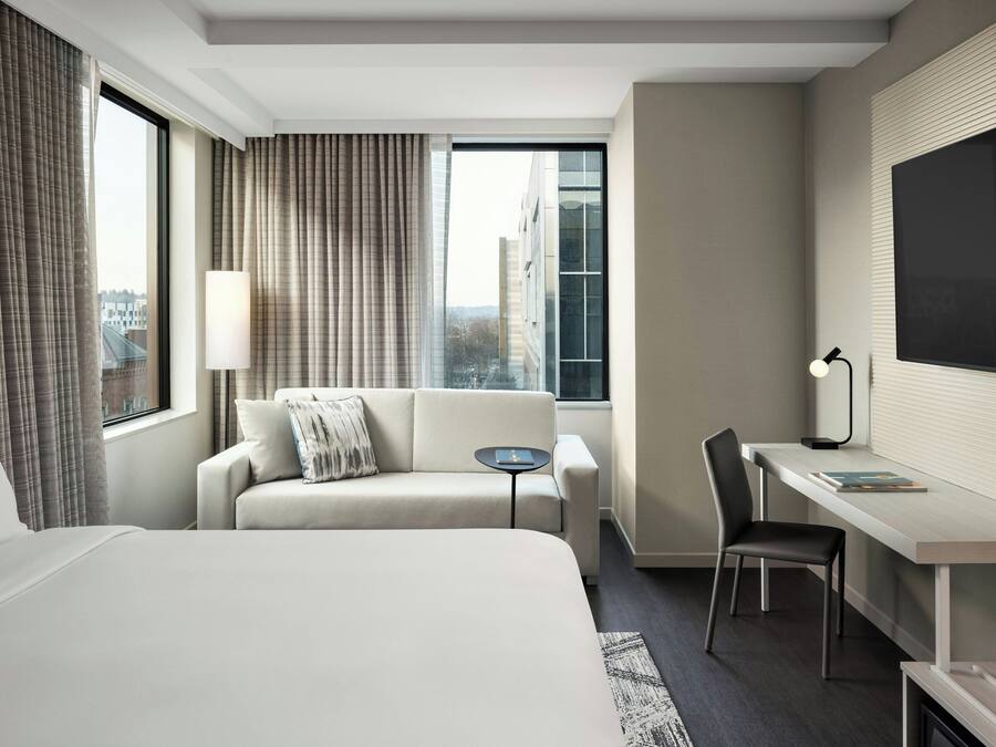 A hotel room with a bed, a sofa, a desk, a chair, a TV, and a window with curtains.