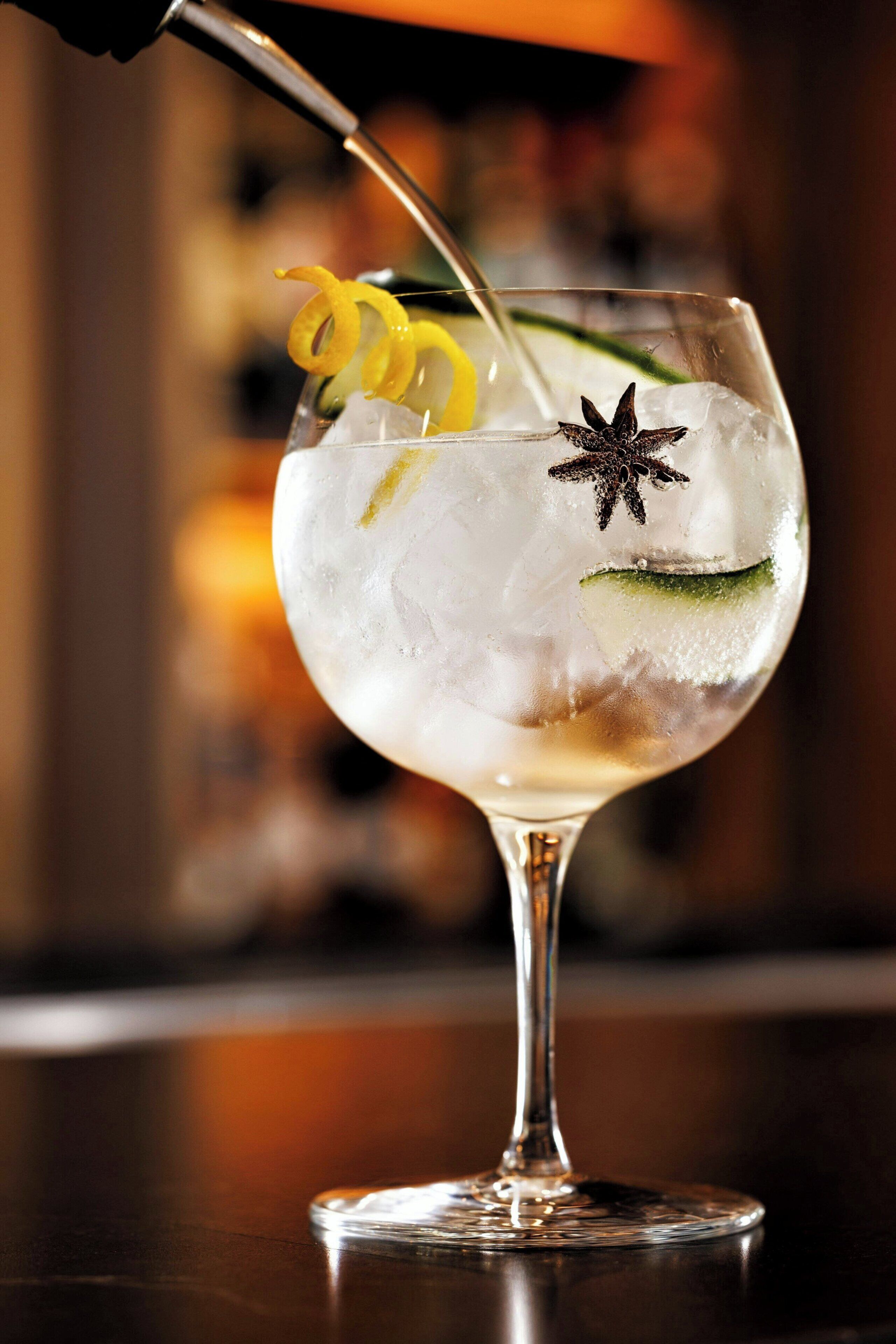 A cocktail being poured into a glass with ice, garnished with a lemon peel and star anise.