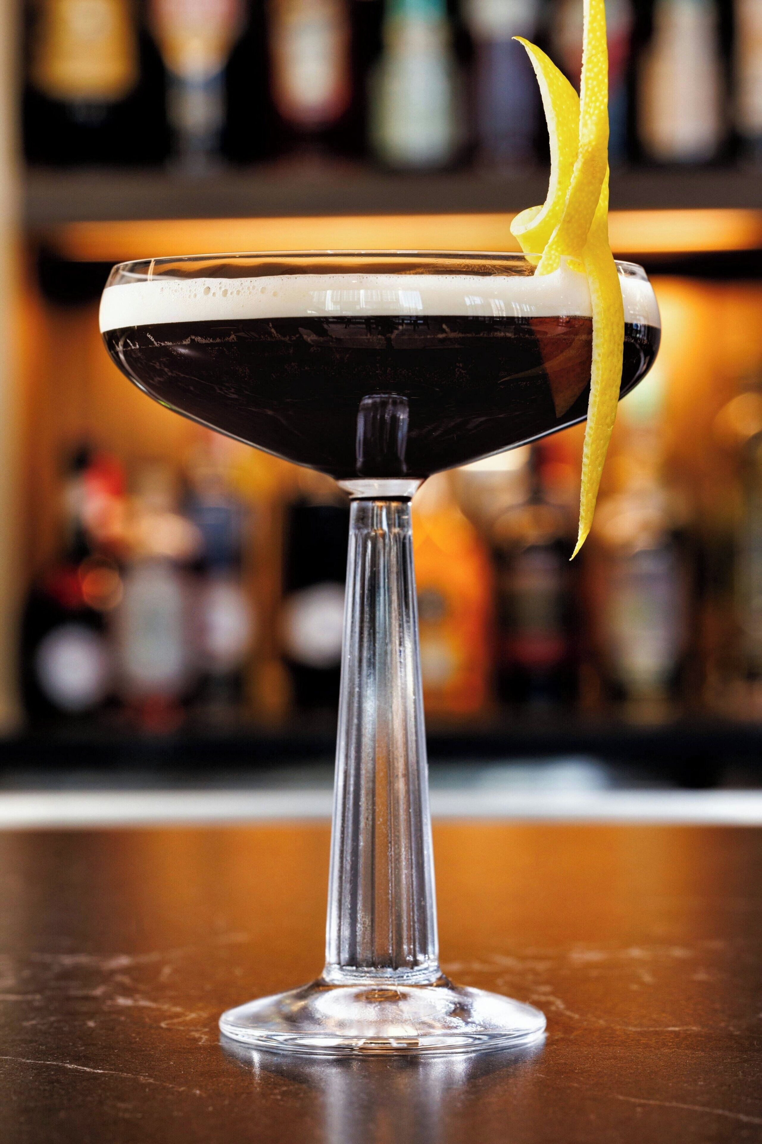 A cocktail in a stemmed glass with a lemon peel garnish, set on a bar counter with a blurred background of bottles.