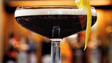 A cocktail in a stemmed glass with a lemon peel garnish, set on a bar counter with a blurred background of bottles.