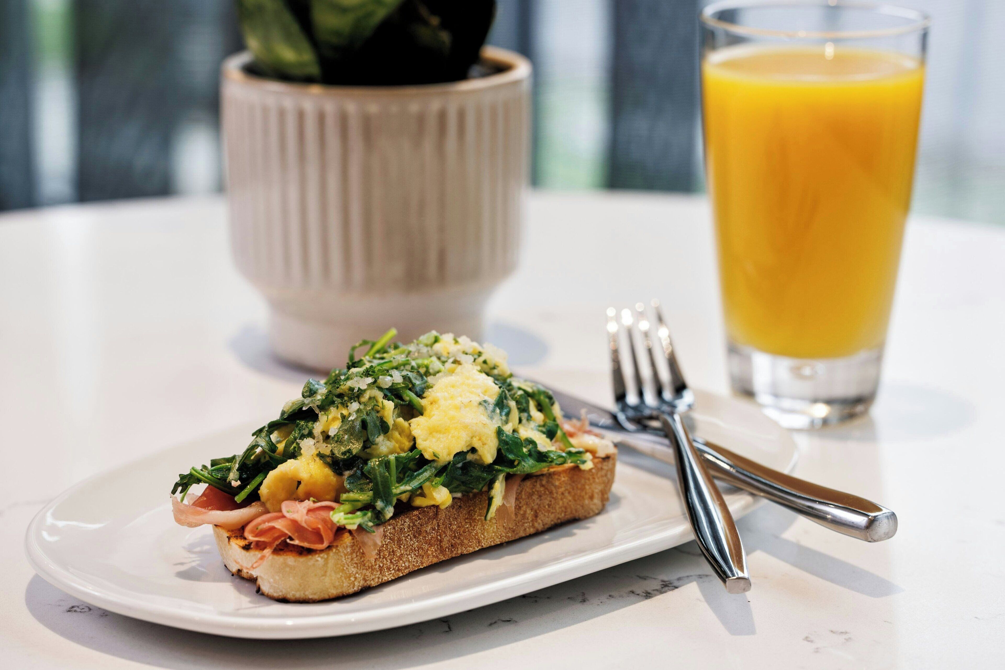 Un plato de desayuno con tostadas, huevos revueltos, verduras, un vaso de zumo de naranja y un tenedor sobre una mesa.