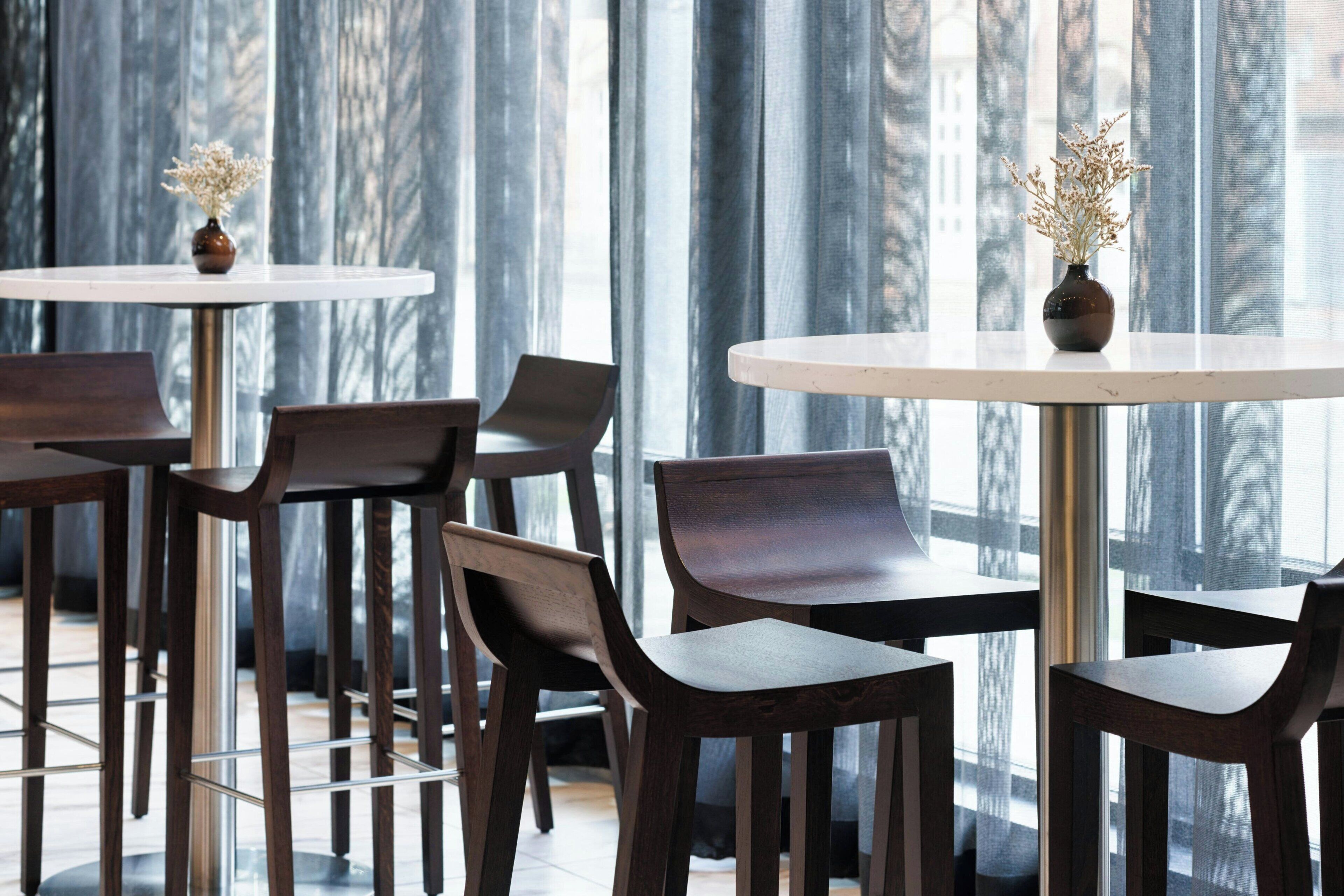 A modern interior with round tables and dark wooden chairs, large windows with vertical blinds, and vases with dried flowers on the tables.