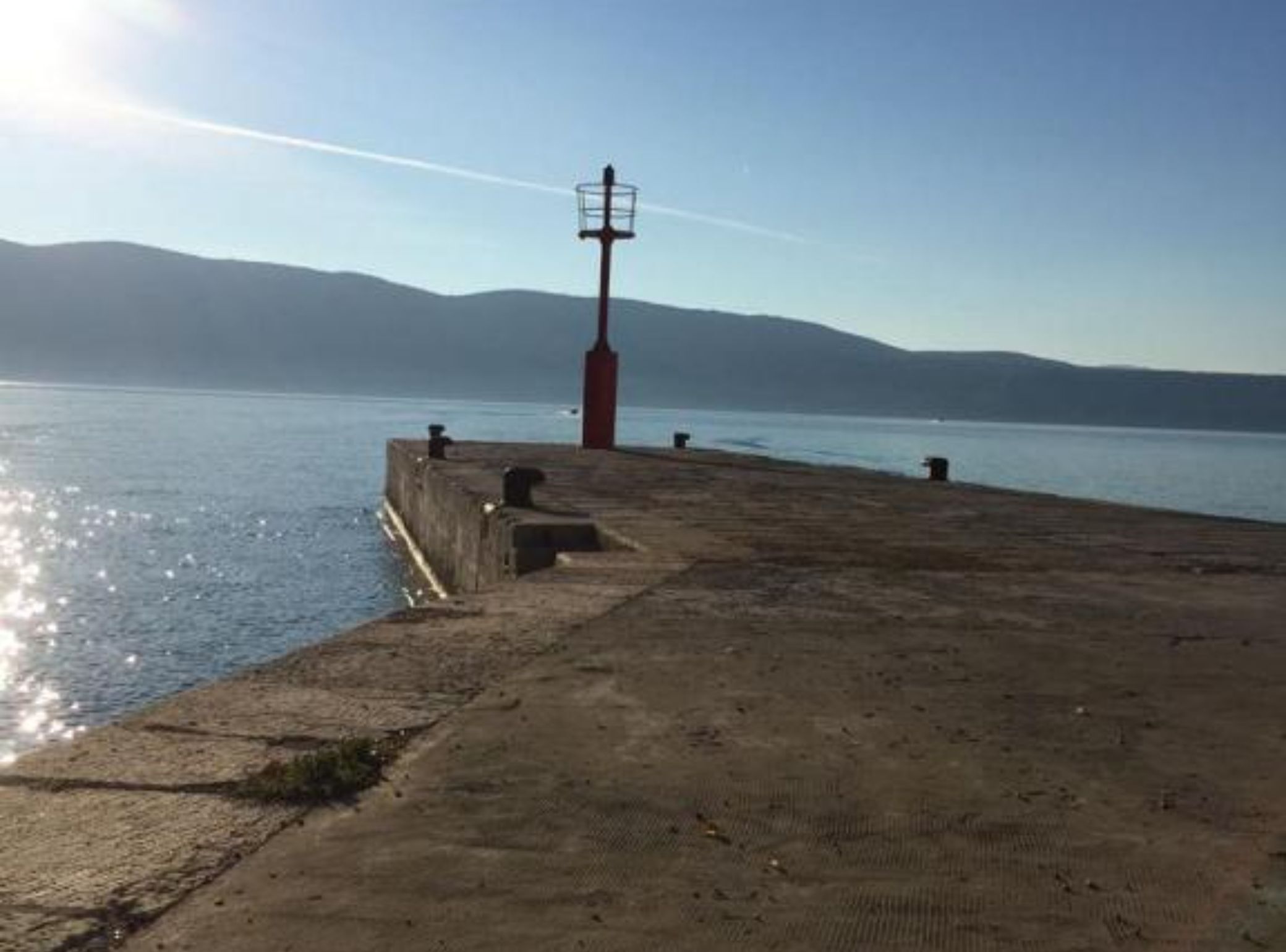 A concrete pier extending into calm water with a red lighthouse in the background.