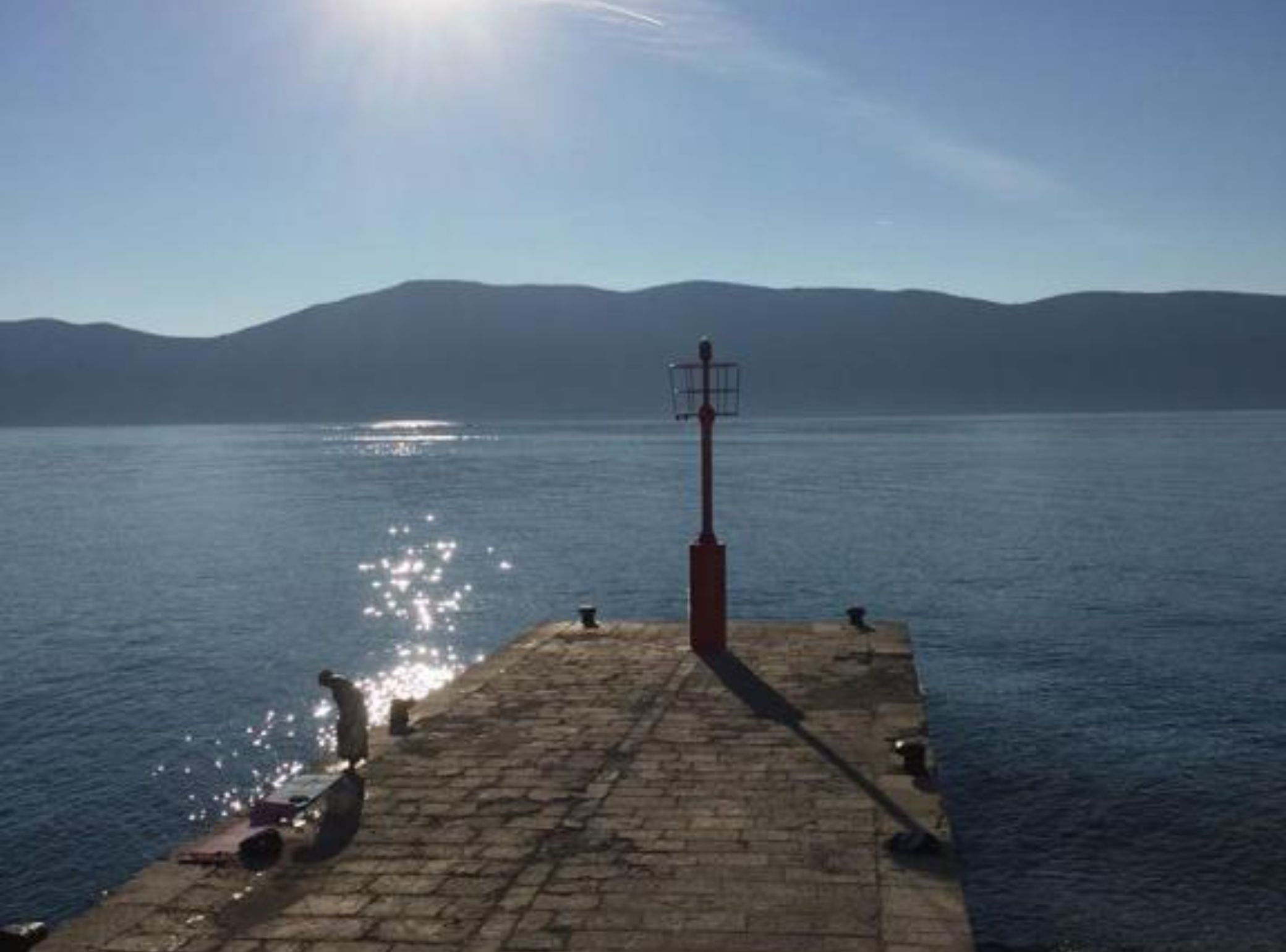Eine steinerne Mole, die sich in ruhiges Wasser erstreckt, mit einem Leuchtturm in der Ferne und Bergen im Hintergrund.