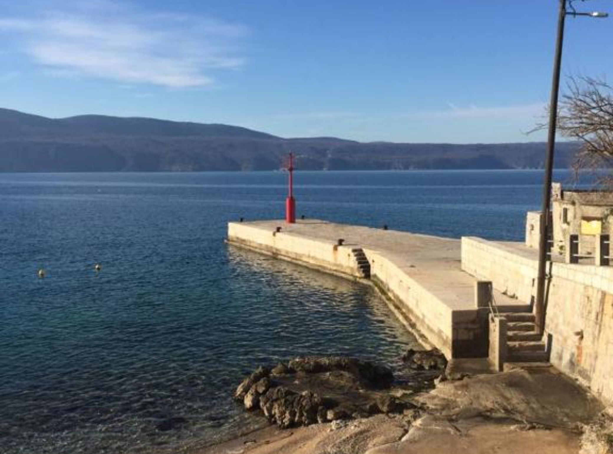 A coastal pier with a red lighthouse, clear water, and distant hills.