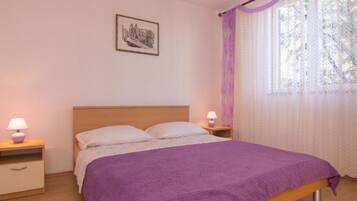 Un dormitorio con una cama de madera, dos mesitas de noche con lámparas, un cuadro enmarcado en la pared y una ventana con cortinas de gasa.