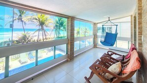 A room with large windows overlooking a beach, a hanging chair, and a wooden bench with cushions. - Condomínio Esmeralda do Mar (Ipojuca)