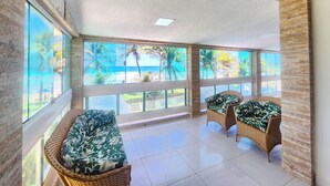 A spacious room with large windows offering a view of the beach and palm trees. There are wicker chairs with green cushions. - Condomínio Esmeralda do Mar (Ipojuca)
