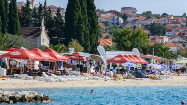 A beach with red umbrellas, a church, and buildings in the background. - Apartments Nela (Okrug)