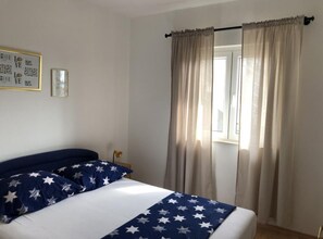 A bedroom with a bed, a window with curtains, a bedside lamp, and a wall with framed pictures. - Apartments Nela (Okrug)