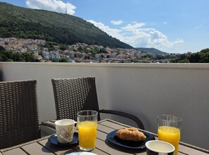 Una mesa con dos sillas de mimbre, dos platos con comida y bebidas, y una vista de un pueblo con casas y una colina al fondo.