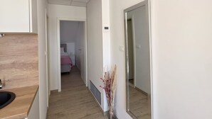 Un pasillo estrecho con suelo de madera, un espejo a la derecha y una puerta que da a un dormitorio con cama y mesita de noche.