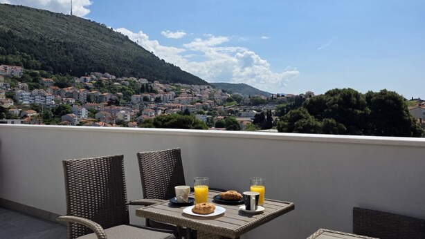 Una terraza con sillones de mimbre y una mesa preparada para el desayuno, con vistas a un pueblo en la colina.