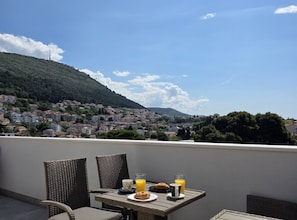 Eine Terrasse mit Korbstühlen und einem für das Frühstück gedeckten Tisch, mit Blick auf eine Hügelstadt.