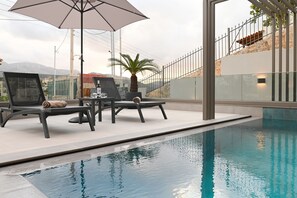 A poolside area with lounge chairs and an umbrella, a palm tree, and a view of a mountainous landscape. - MGK Complex Suite Private Pool Ground Floor (Malevizi)