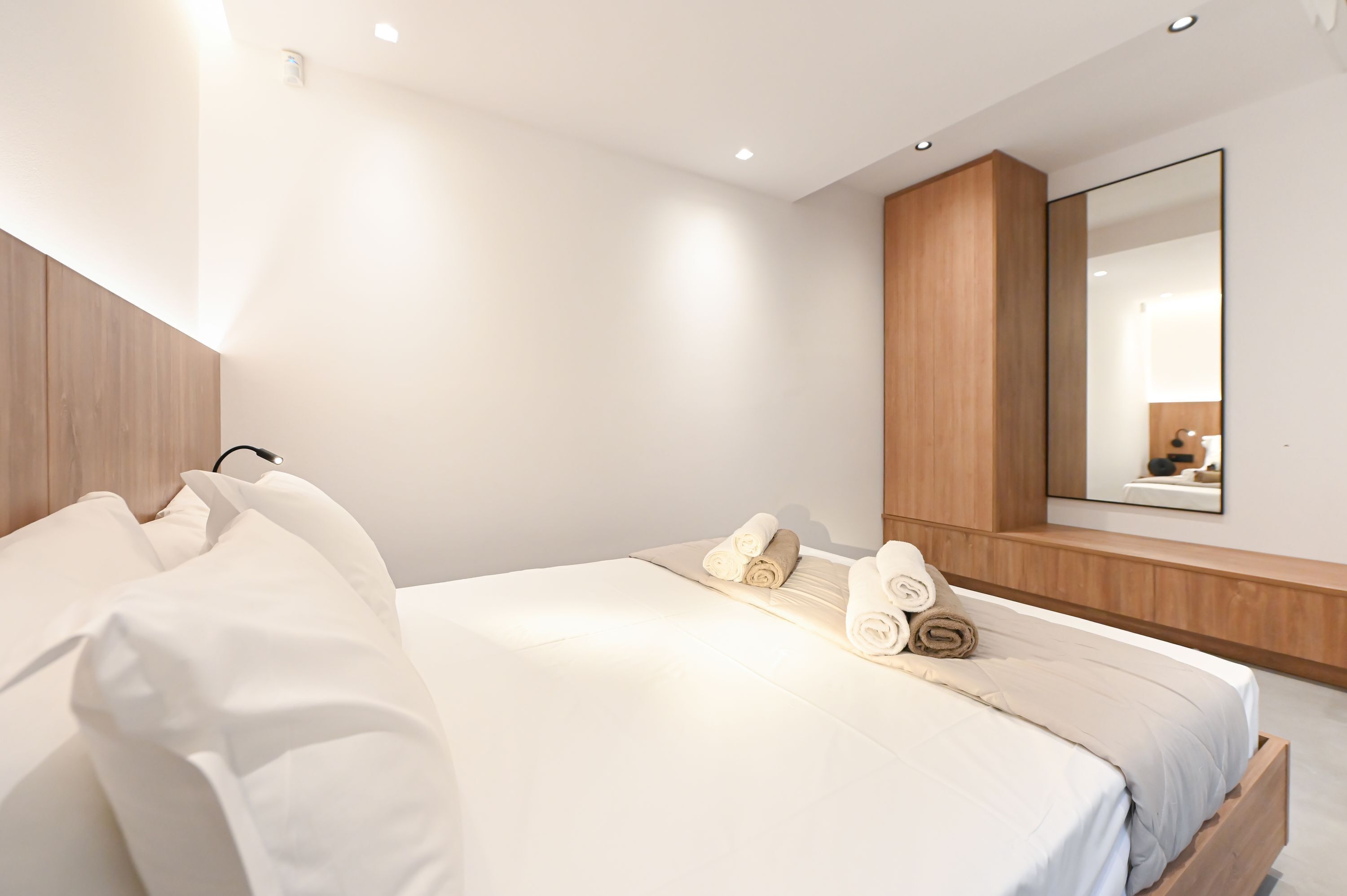 A modern hotel room with a large bed, wooden headboard, a built-in bench, and a large mirror.