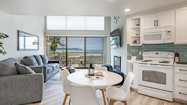 A modern kitchen with white cabinets, a built-in microwave, and a stove. There is a dining area with a round table and chairs, and a gray sofa in the living room.