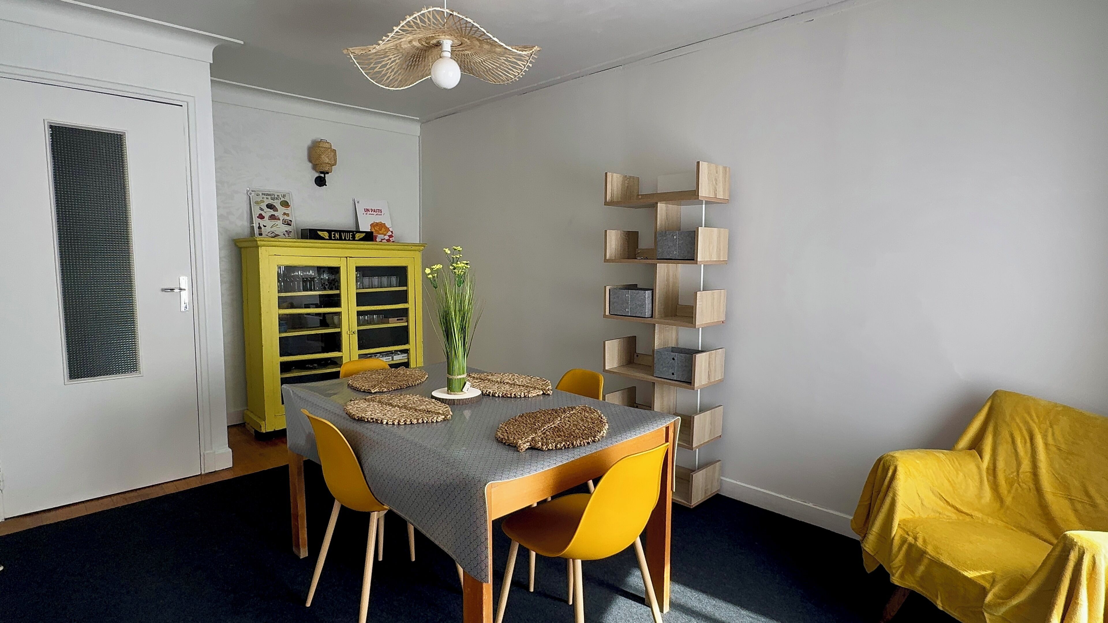 A dining area with a table set for four, yellow chairs, a yellow cabinet with glass doors, and a wall-mounted wooden shelf unit.